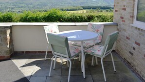 A patio with a table and chairs, a brick wall, and a view of green hills. - 2 bedroom accommodation in Llanrhaeadr, near Denbigh (Llanrhaeadr, near Denbigh)