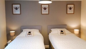 A hotel room with two beds, each with a bee-themed pillow, a nightstand with a lamp, and framed bee artwork on the wall. - 2 bedroom accommodation in Llanrhaeadr, near Denbigh (Llanrhaeadr, near Denbigh)