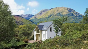 Une petite maison blanche, dotée d’un toit en pente et d’un escalier menant à l’entrée, est entourée d’une végétation luxuriante et, au loin, on aperçoit des montagnes.