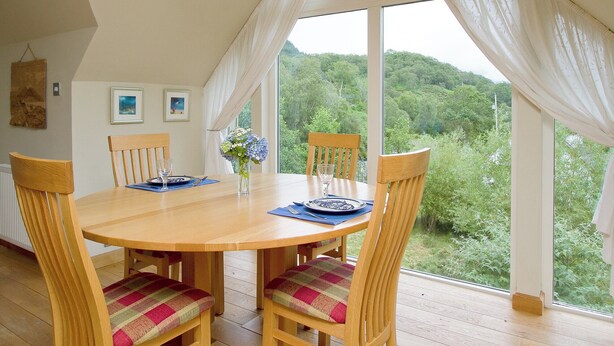Un coin repas comprenant une table en bois, quatre chaises, un lustre et de grandes fenêtres avec des rideaux.