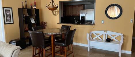 A dining area with a round table and chairs, a white bench, and a kitchen with a stainless steel refrigerator.