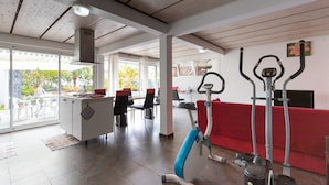 Une pièce spacieuse équipée d’appareils de musculation, d’un coin repas et de grandes fenêtres donnant sur l’extérieur.