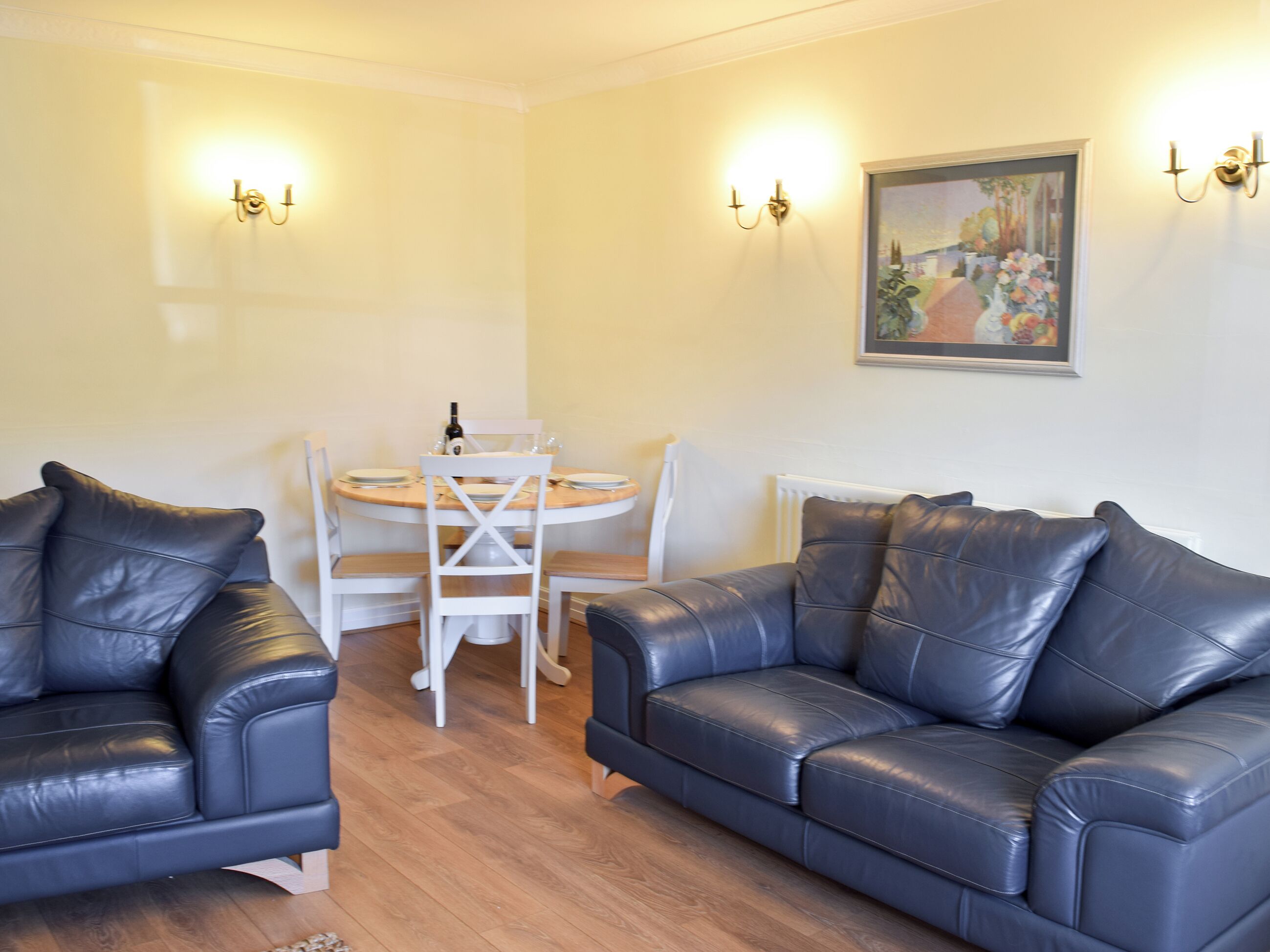A living room with two black leather sofas, a round wooden table with chairs, and wall-mounted lighting.