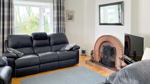 A modern living room with a black leather sofa, a grey rug, a brick fireplace, and a television. - 3 bedroom accommodation in Bittaford, near Ivybridge (Bittaford, near Ivybridge)
