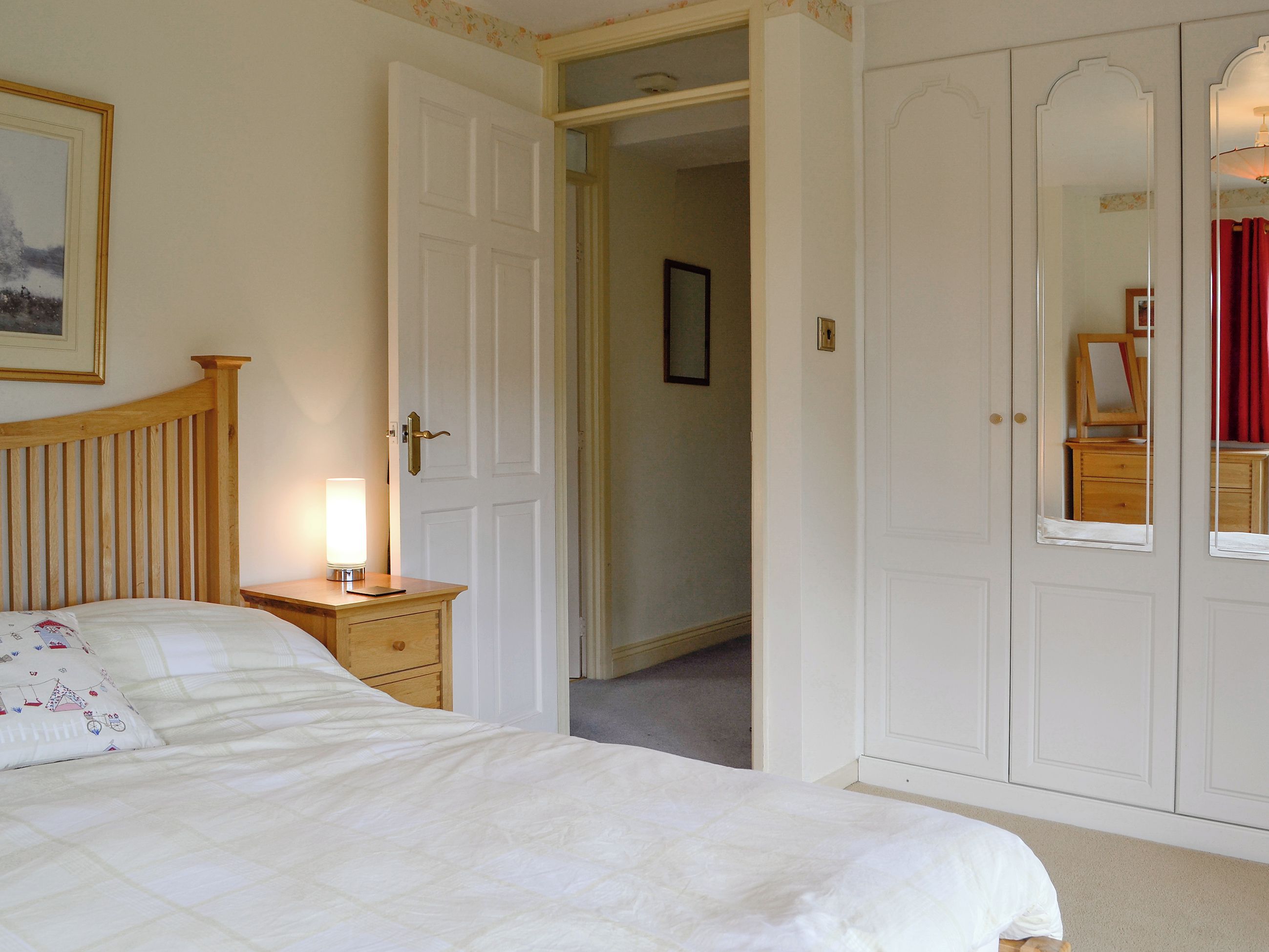A bedroom with a bed, bedside table, lamp, wardrobe, and a door leading to another room.