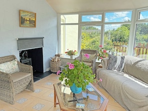 A sunlit room with a fireplace, wicker furniture, a glass coffee table, and a view of greenery outside.