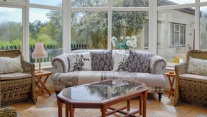 Een glazen orangerie met een bank, twee rieten fauteuils, een glazen tafel en een lamp.