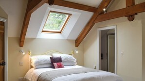 Une chambre avec un plafond en pente, une lucarne, un lit avec une literie blanche et une structure de poutres apparentes en bois au plafond.