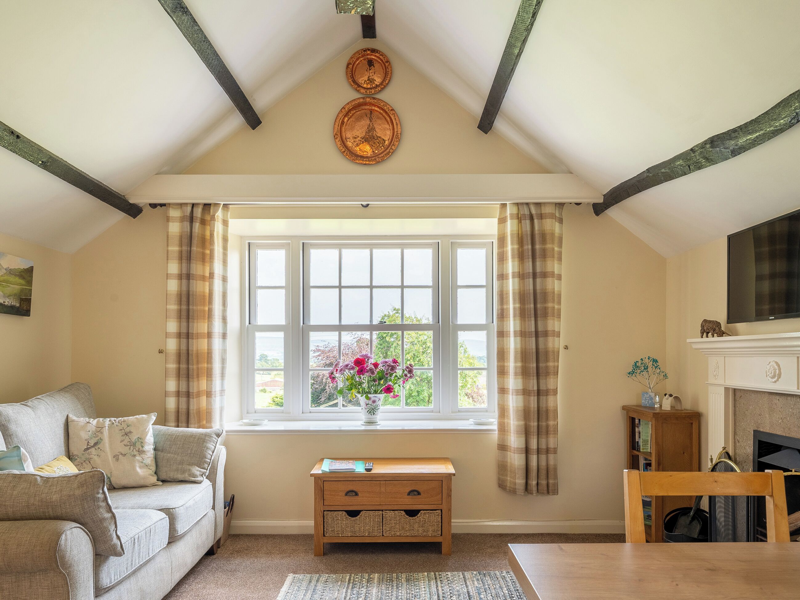 A cozy living room with a sofa, a wooden side table, a fireplace, and a large window with curtains.