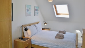 Un dormitorio con techo inclinado, una cama con cabecera, una mesita de noche con lámpara, otro mesita auxiliar y dos obras de arte en la pared.