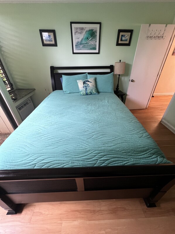 A bed with a teal quilt and a decorative pillow, a nightstand with a lamp, and framed pictures on the wall.