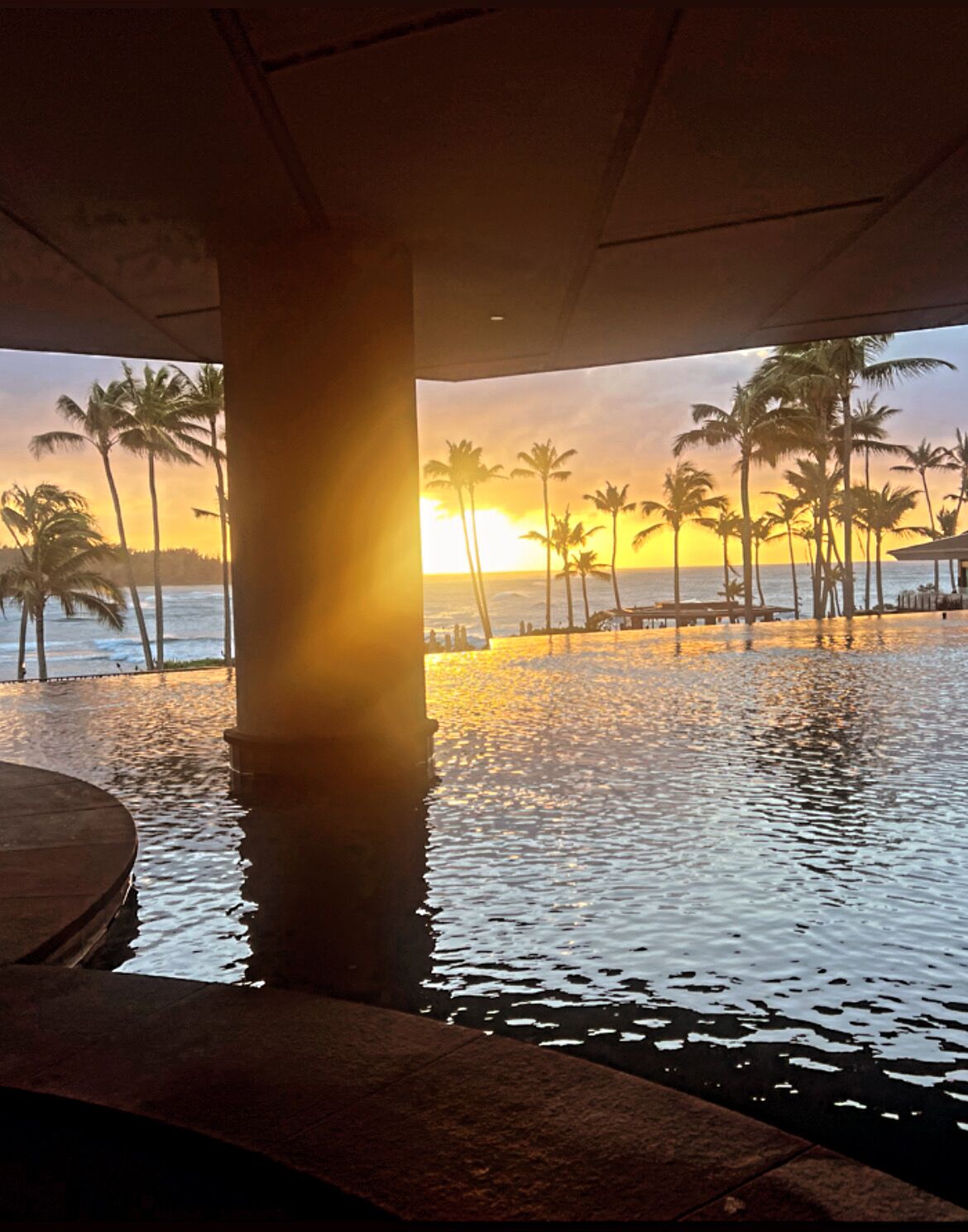 Blick auf einen Strand mit Palmen und einem Sonnenuntergang über dem Wasser, aufgenommen aus einer Struktur heraus.