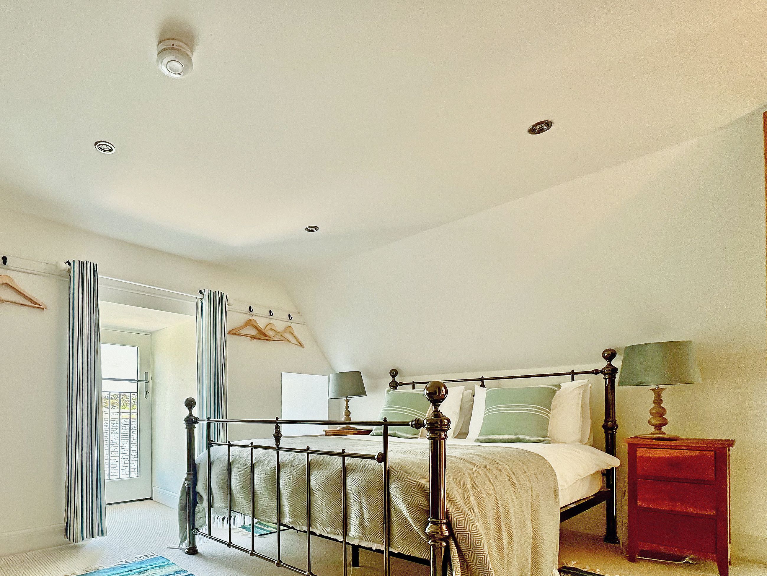 Une chambre à coucher comprenant un lit, des tables de chevet, un porte-manteau mural et une porte donnant sur l’extérieur.