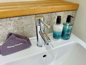 Un lavamanos con grifo cromado, una toalla morada con la etiqueta 'Desmaquillante' y dos botellas de jabón de manos y cuerpo 'Scottish Fine Soaps'.