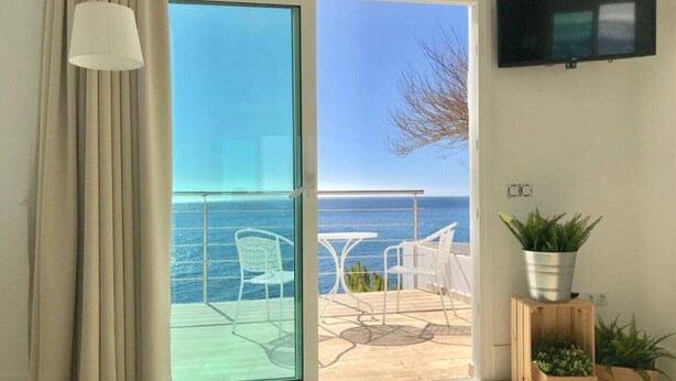 Ett rum med en skjutdörr i glas som leder ut till en balkong med utsikt över havet. På balkongen finns vita stolar och ett bord, och en tv är monterad på väggen.