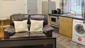 A compact living space with a brown leather sofa, a small kitchen area including a microwave and a washing machine, and a window with curtains. - 1 bedroom accommodation in Aberdeen (Aberdeen)
