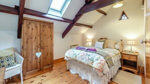 A bedroom with a sloped ceiling, wooden beams, a large wardrobe, a bed with floral bedding, bedside lamps, and a small window. - 1 bedroom accommodation in Gowerton, Gower (Gowerton, Gower)