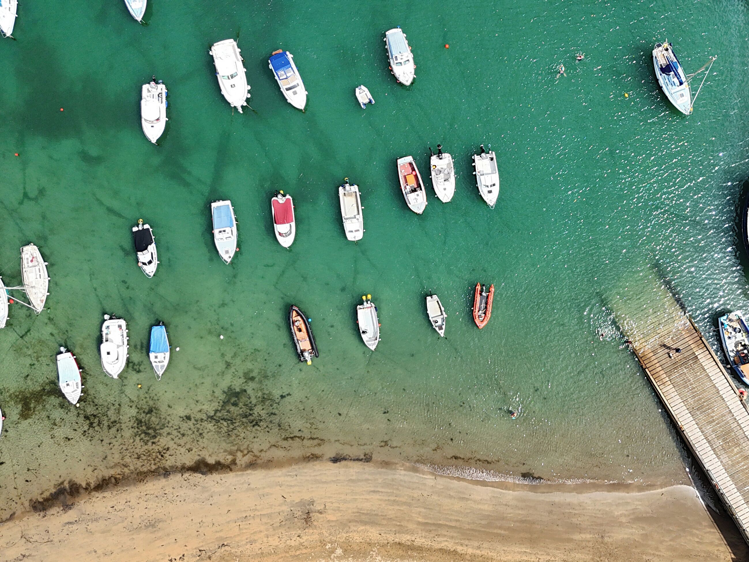 An aerial view of a marina with various boats docked, a sandy beach, and a wooden pier.