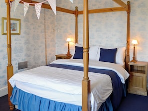 A four-poster bed with a wooden frame, white and blue bedding, two bedside tables with lamps, and a patterned wallpaper background.
