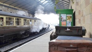 Ein alter Zug an einem Bahnhof, aus dessen Motor Dampf aufsteigt.