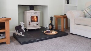 Un salon chaleureux avec un poêle à bois blanc, un canapé blanc et une petite table en bois sur laquelle se trouvent une bouteille et des raisins.