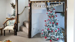 Une pièce chaleureuse avec un sapin de Noël, une chaise en bois, un ventilateur et une petite table sur laquelle se trouvent des fruits.
