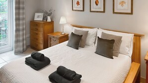 Una cama bien hecha con almohadas y toallas dobladas, un aparador de madera, una mesita de noche con lámpara y una obra de arte con hojas enmarcadas en la pared.