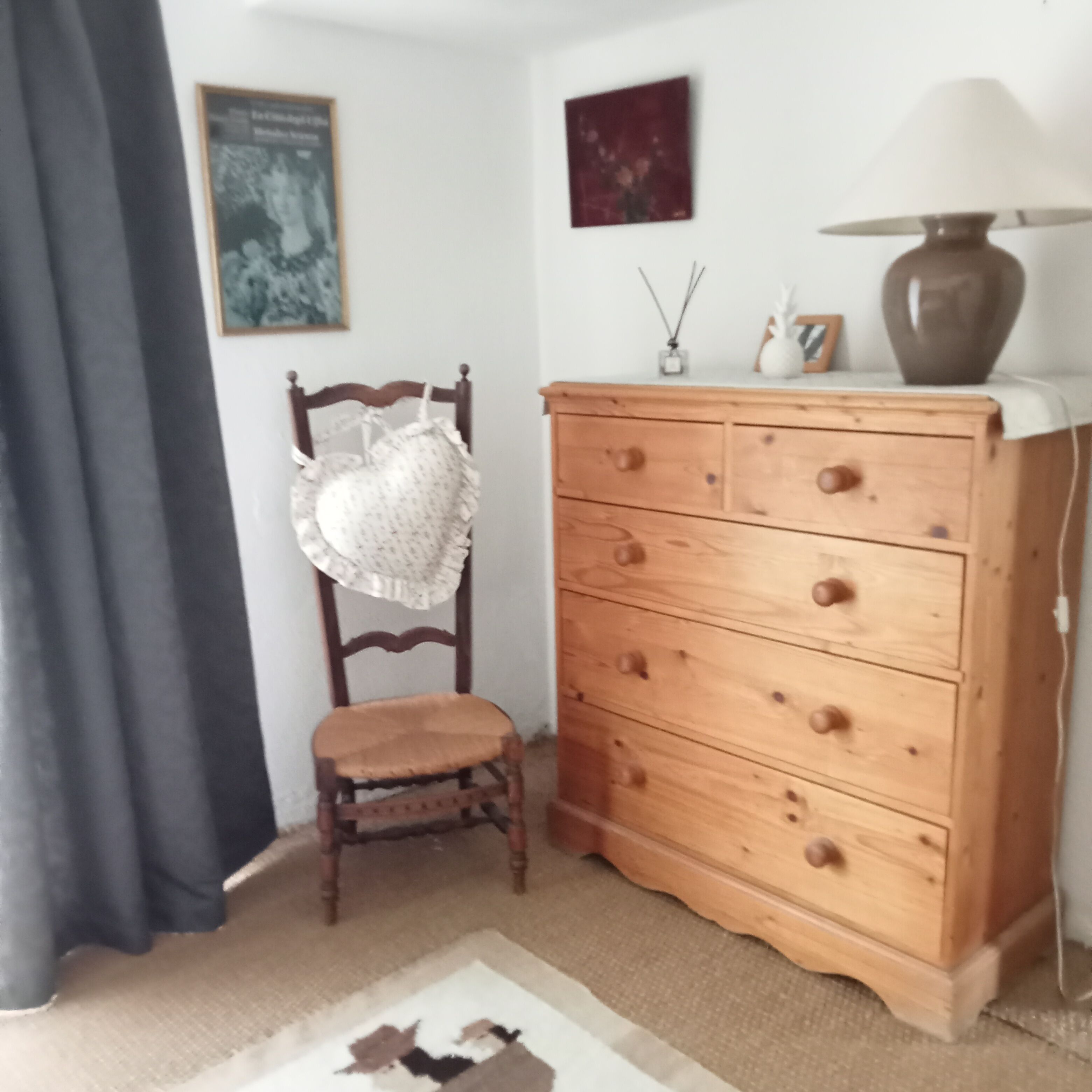 Ein Zimmer mit einem Holzkleiderschrank, einem Sessel mit Kissen mit Herzmuster, einem gerahmten Bild an der Wand und einer Lampe auf dem Schrank.