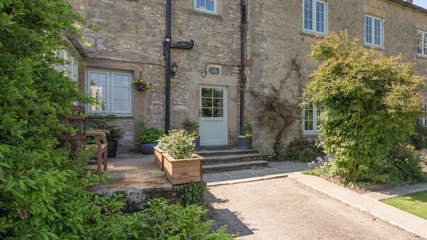 A stone building with a wooden door, windows, and a small garden area with potted plants.