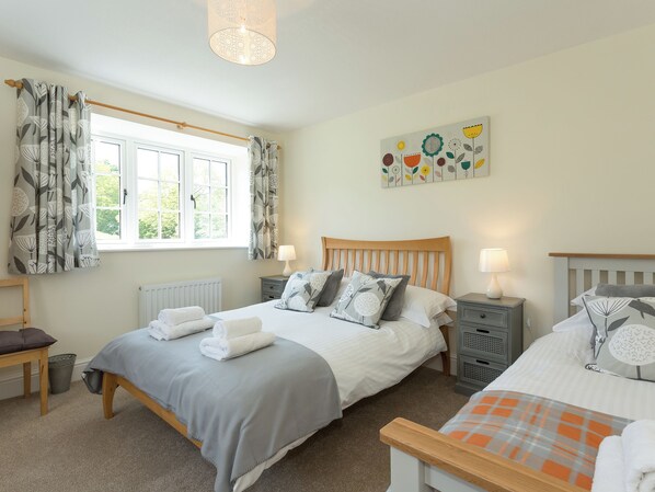 A bedroom with two beds, a window with curtains, a radiator, a wooden headboard, bedside tables with lamps, and a floral artwork on the wall.