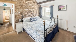 A bedroom with a stone wall, a bed with a blue and white patterned bedspread, a wooden headboard, a nightstand, a window with curtains, and a radiator. - 1 bedroom accommodation in North Stainmore, Kirkby Stephen (North Stainmore, Kirkby Stephen)