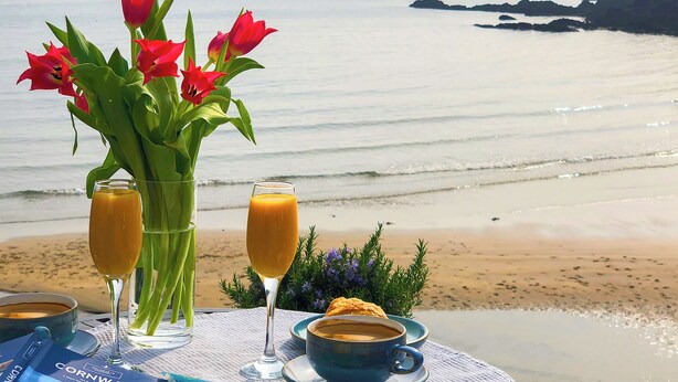 A table with a view of the sea, a bouquet of red flowers, and a book titled "Cornwall". - Puffin Burrow, a beautiful historic couples retreat overlooking the Cove. (Portmellon, near Mevagissey)