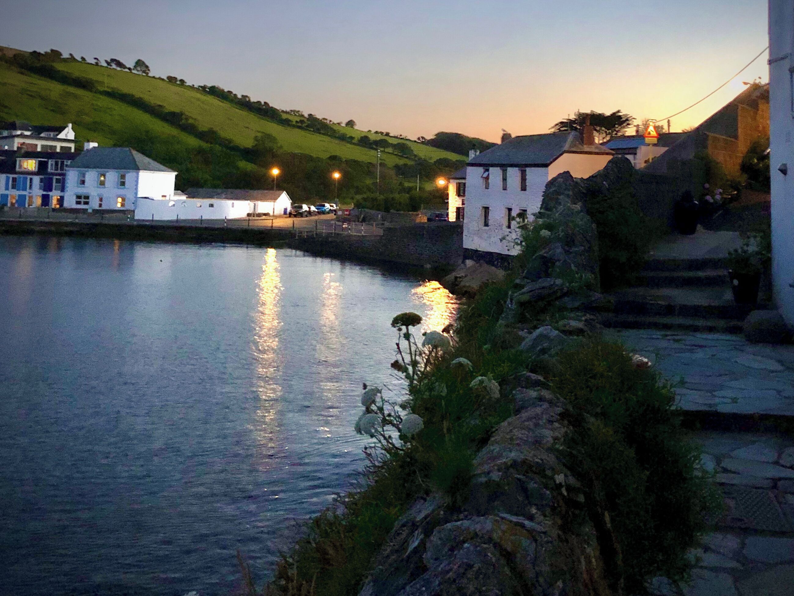 A coastal town at dusk with buildings along the water, a stone pathway, and a hill in the background.