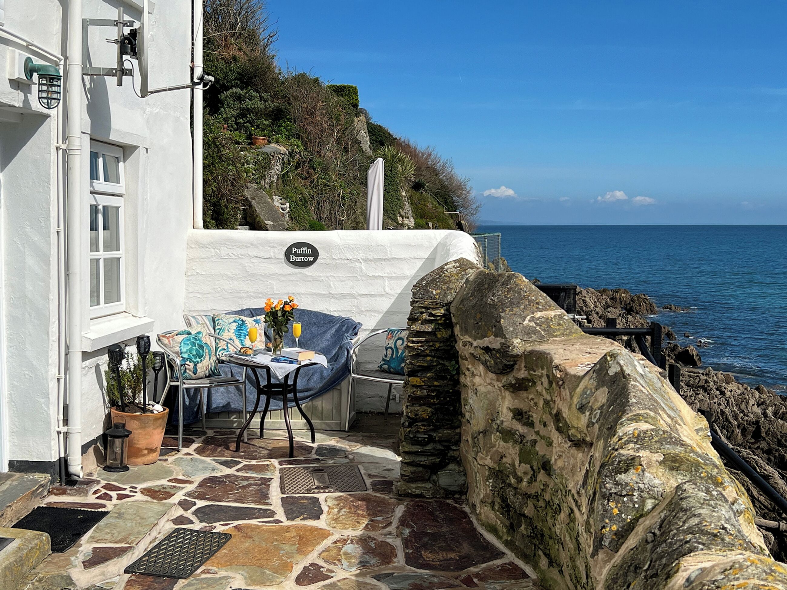 A stone patio with a table and chairs, a potted plant, and a view of the sea.