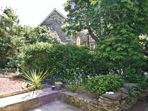 A stone house with a garden in front, including a stone wall, potted plants, and flowering shrubs.