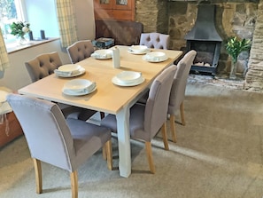 Un coin repas avec une table en bois dressée pour six personnes, entourée de chaises rembourrées. On aperçoit une cheminée en pierre au fond.