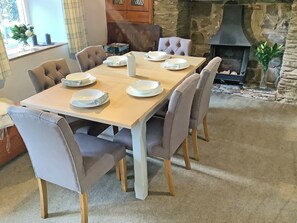 A dining area with a wooden table set for six, surrounded by upholstered chairs. A stone fireplace is visible in the background.