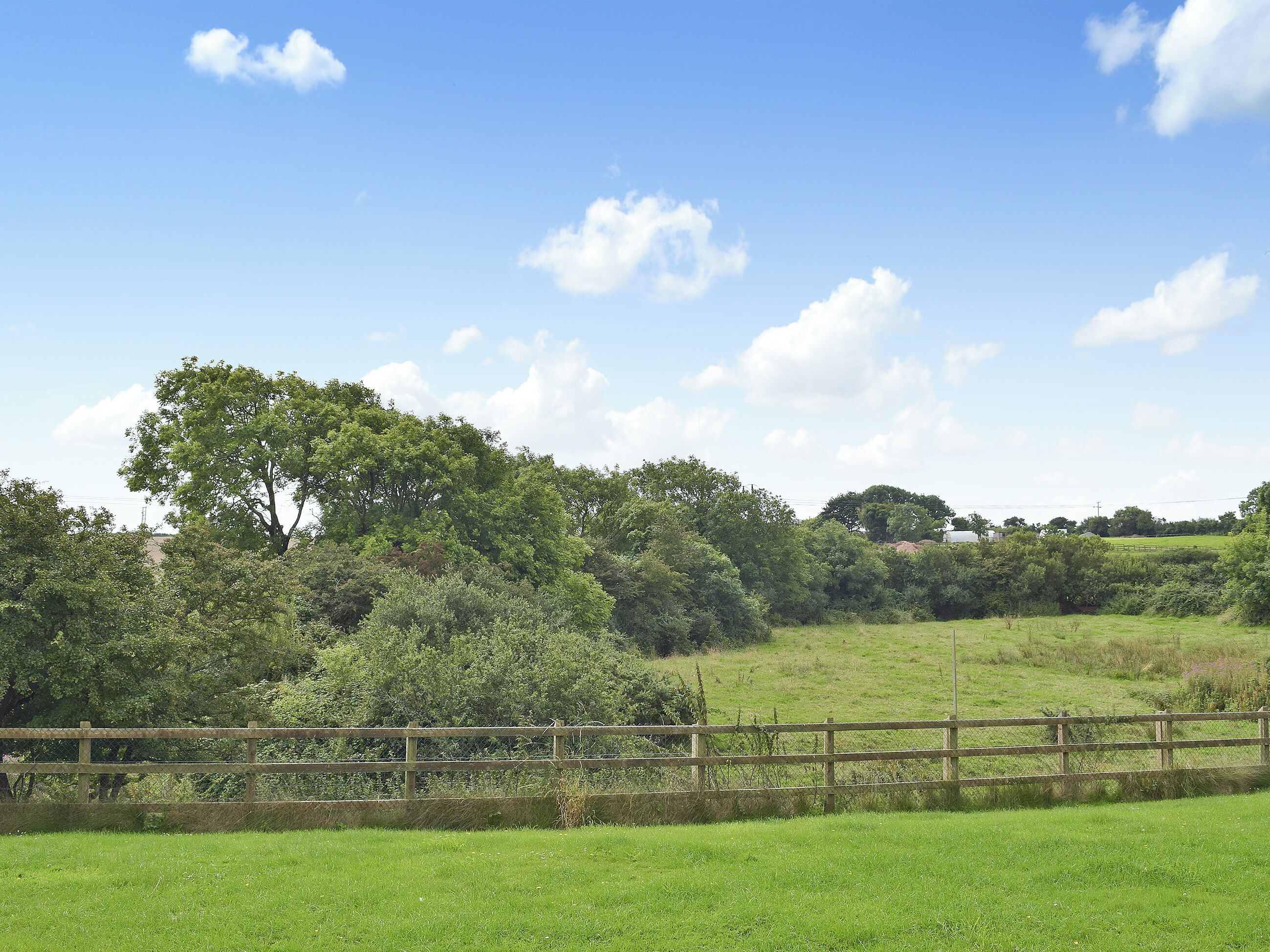 A rural landscape with a wooden fence, lush green fields, and dense trees under a clear blue sky.