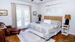 Ein Schlafzimmer mit einem Bett, zwei Nachttischen, einem Sessel, einem Schreibtisch, einem Fenster mit Vorhängen und einem Deckenventilator.