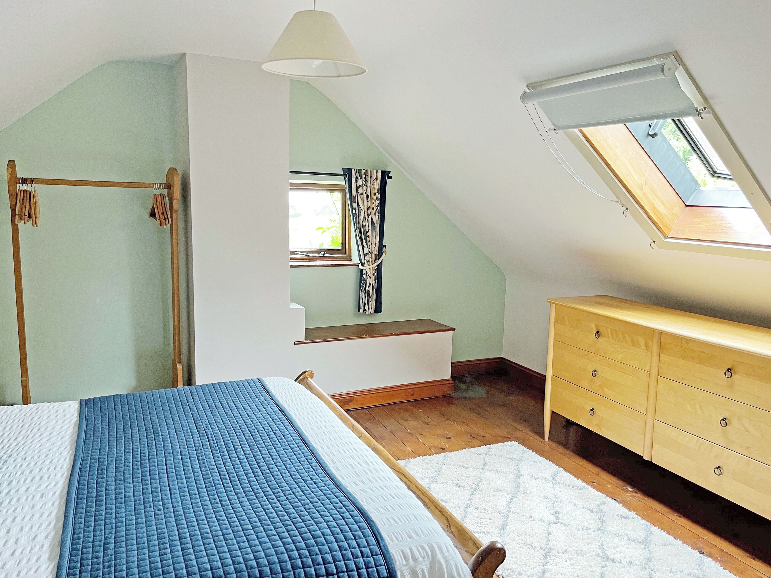 Un dormitorio con techo inclinado, una cama con colcha azul, un aparador de madera y un tragaluz.