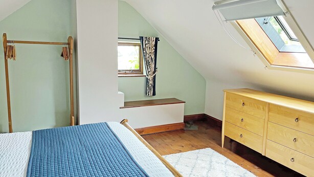 Une chambre avec un plafond en pente, un lit recouvert d’une courtepointe bleue, une commode en bois et un puits de lumière.