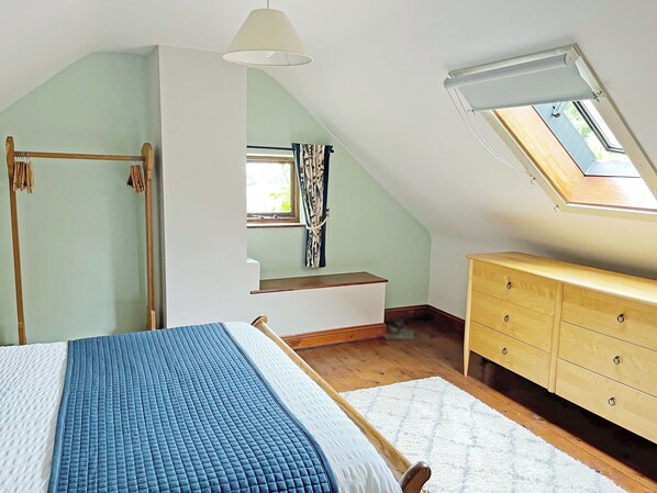 Ein Schlafzimmer mit schrägen Wänden, einem Bett mit blauer Steppdecke, einer hölzernen Kommode und einem Dachfenster.