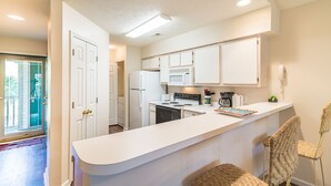 A modern kitchen with white cabinetry, a central island, and a dining area with wicker chairs. - TB 9I Super 3rd floor condo True Blue Golf/Tennis Accessible WINTER RENTAL (Pawleys Island)