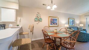A dining area with a table and chairs, a kitchen area with a coffee maker, and a living area with a couch and a lamp. - TB 9I Super 3rd floor condo True Blue Golf/Tennis Accessible WINTER RENTAL (Pawleys Island)