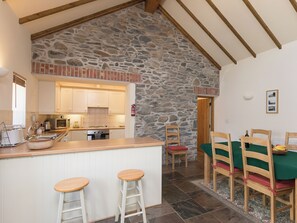 A kitchen with a stone wall, wooden beams, a dining area with a green tablecloth, and a bar with stools.