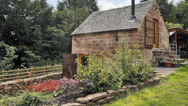 A stone cottage with a sloped roof, a chimney, and a garden with various plants and a wooden fence.