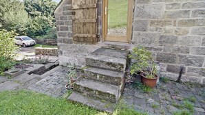 A stone cottage with a wooden door and shutter, a small set of stone steps, and a car parked outside.