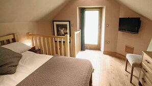 A bedroom with a sloped ceiling, a bed with a headboard, a wooden chair, a dresser, a television mounted on the wall, and a framed picture on the wall.