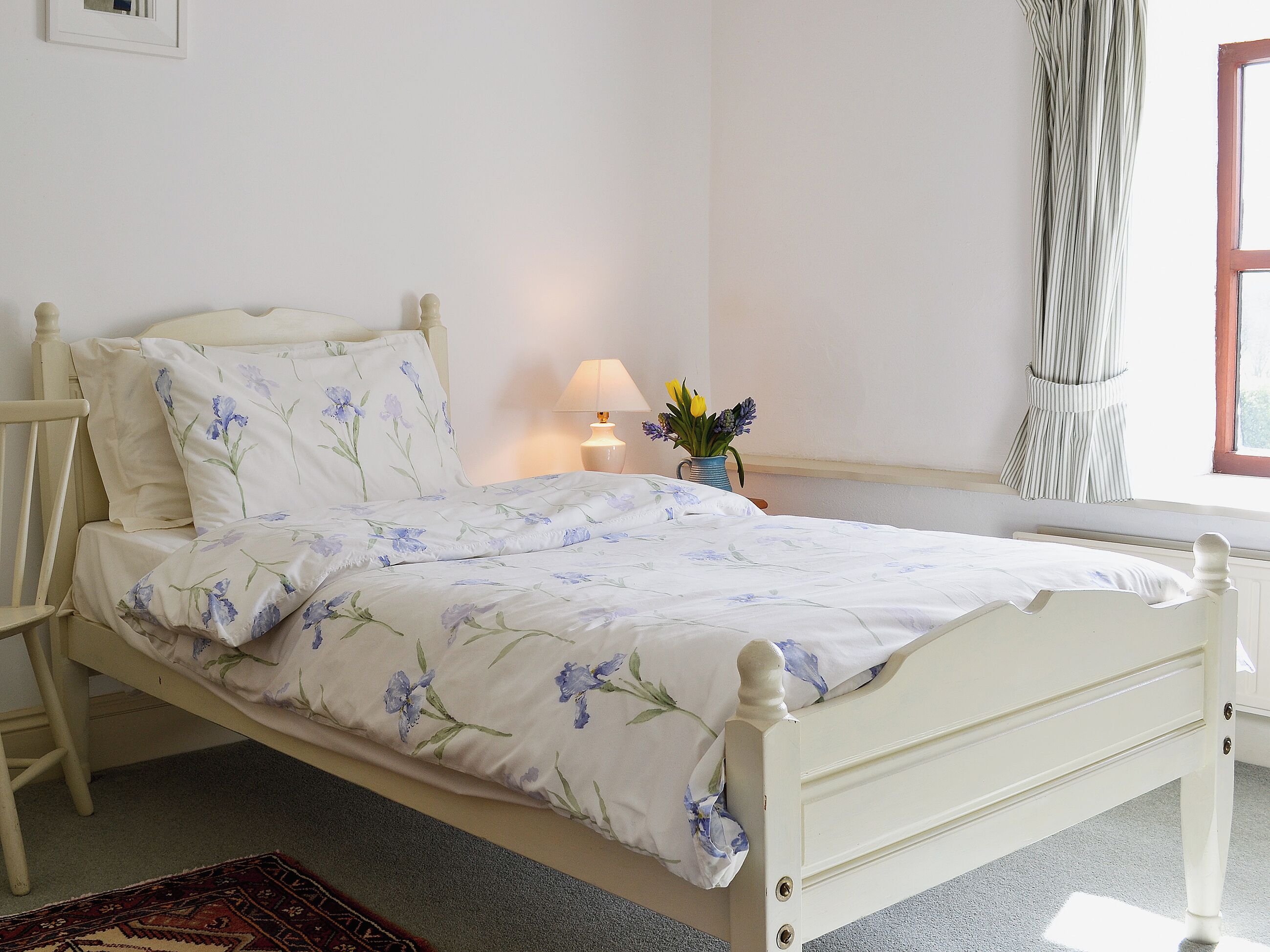 A neatly made bed with floral bedding, a bedside lamp, and a vase with flowers on a nightstand.