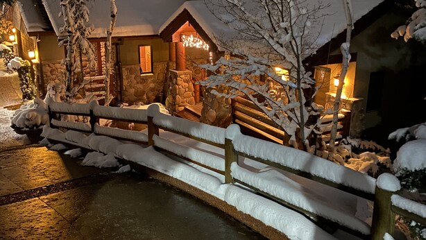 Un refugi cobert de neu amb façana de pedra, un pont de fusta i un interior il·luminat visible a través de les finestres.